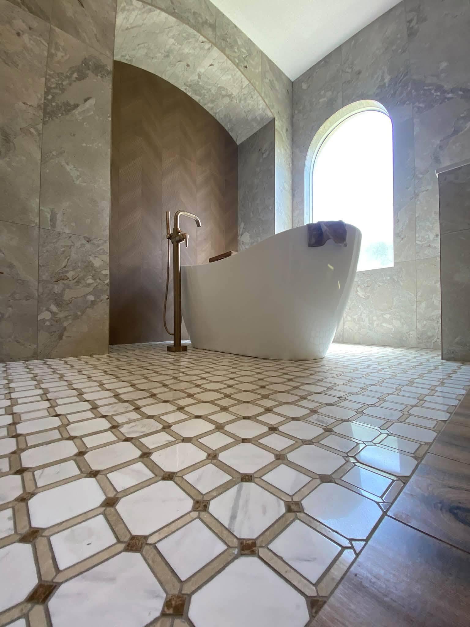 Freestanding white tub standing in an arched alcove lined with chevron-textured tile, marble-look walls either side, an arched window to the right and a geometric mosaic floor running to a wood floor edge