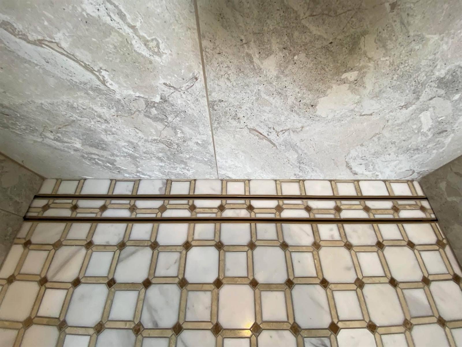 Linear shower drain set flush into an octagon mosaic floor with brass-toned inserts, running tight against a marble-look wall