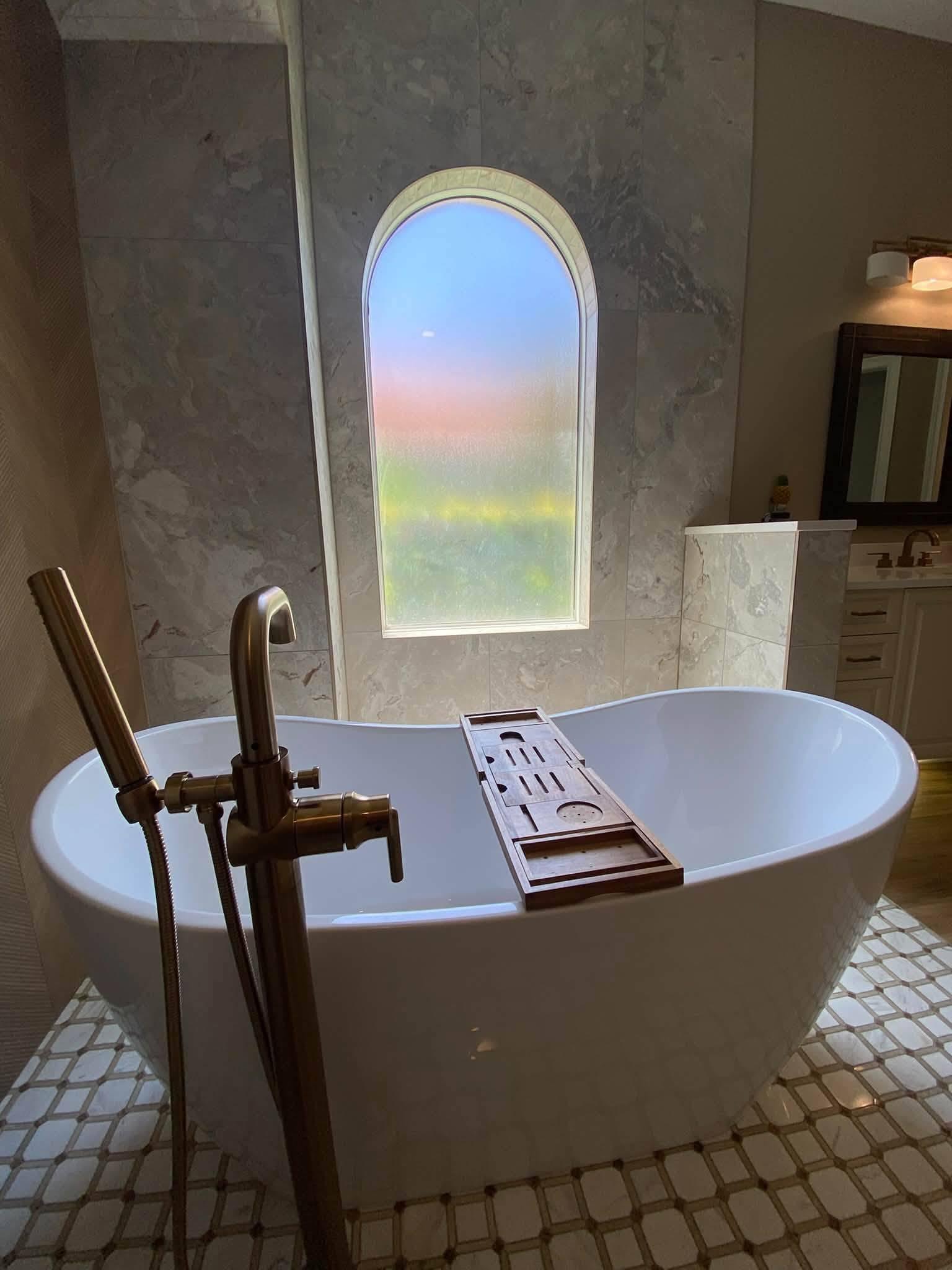 The freestanding tub in evening light with a brass floor-mounted filler, an arched window behind it and the vanity visible beyond