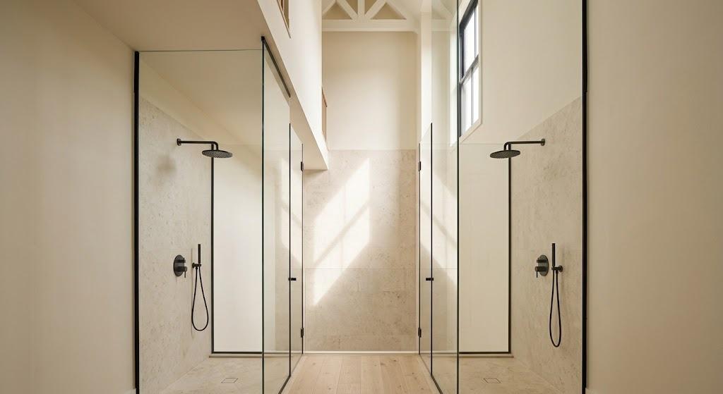 Double height glass enclosed shower in pale stone tile with two rain heads on opposite walls and a tall window high up throwing daylight down the wall (illustrative rendering, not a photo of the project)